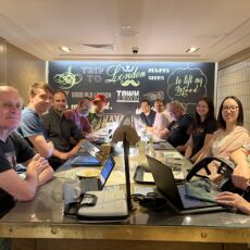 11 attendees sitting round a long table in cafe with laptops and the backdrop of a mural-style decorated wall with Trip to London Town