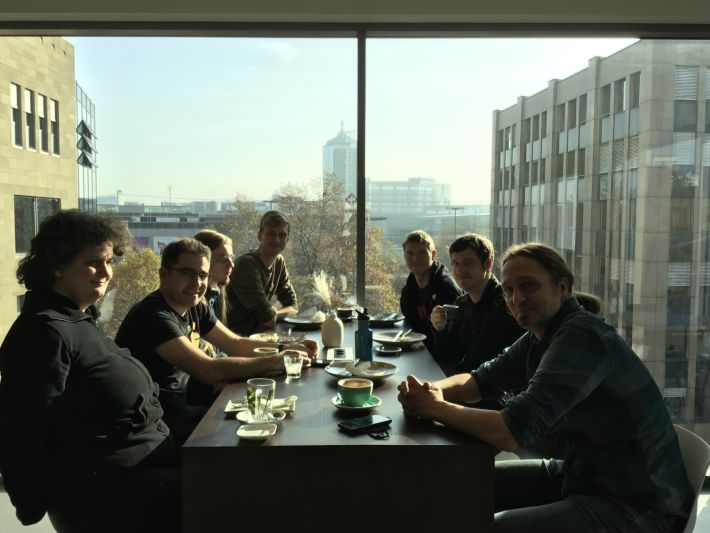 IndieWebCamp Düsseldorf folks sitting at a table in front of a window for lunch.