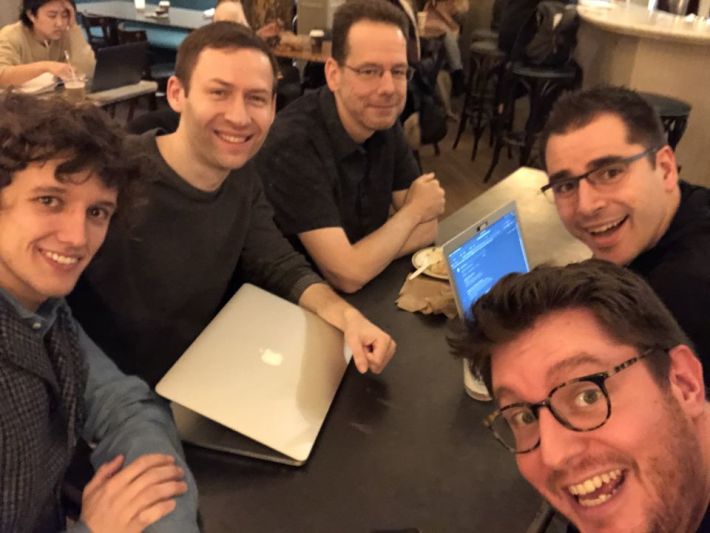 5 attendees smile at the camera while huddling around a metal table with two laptops visible.