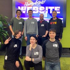 Participants of HWC San Francisco pose in two rows in front of a large screen displaying an 80s style Homebrew Website Club logo in the commons room at Mozilla San Francisco.