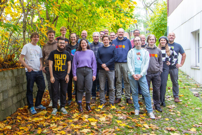 Twenty IndieWebCamp participants standing smiling roughly in 3-4 rows on an outdoors inclined driveway with yellow leaves and grass on the ground, and a canopy of trees overhead, between a mid-height retaining wall and building.
