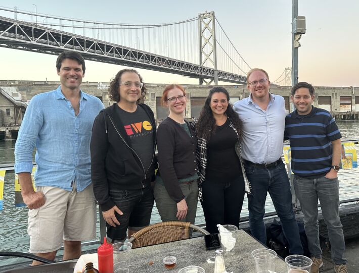 Six people standing side-by-side near an outside dining table at Red’s Java House with the Bay Bridge in the background just after sunset.