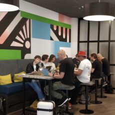 Several people using laptops sitting at a table in a spacious lobby with a large abstract artwork on the wall behind them