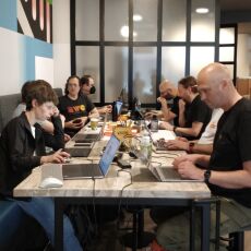 Nine people sitting with laptops at a high table, stools on one side, long couch on the other, typing and having discussions.