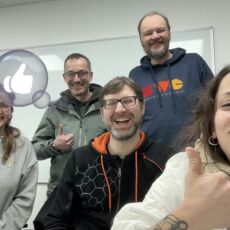 Five people smiling, sitting and standing staggered in front of a white board, with a semi-transparent graphic thumbs-up in a bubble seemingly originating from a thumbs-up gesture of one of the people.