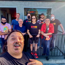 Man taking a selfie in the foreground, with 8 people standing in front of a storefront in the background.