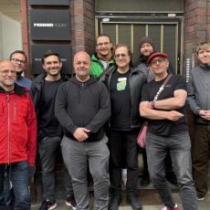 IndieWebCamp participants standing up overlapping in front of a brick building with a recessed door.