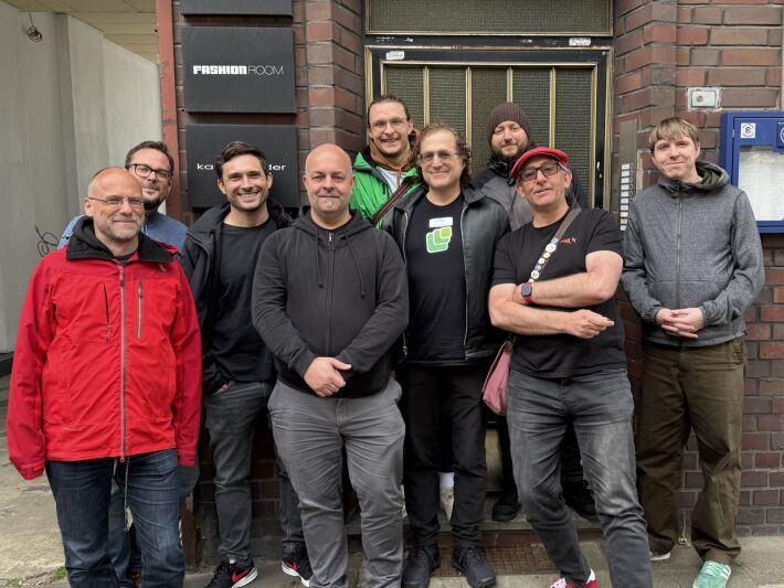 IndieWebCamp participants standing up overlapping in front of a brick building with a recessed door.