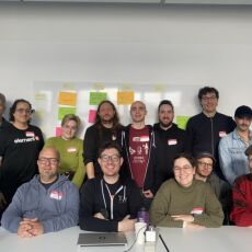 14 of the participants at IndieWebCamp Berlin lined up in two rows, one sitting, one standing in back, with part of the breakouts schedule grid behind them, white tables in front and a white wall behind them, well lit by natural light.
