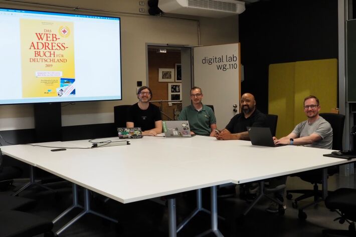 Four people sit at a conference table in a modern meeting room called "digital lab wg.110," with laptops and a large screen displaying the cover of "Das Web-Adressbuch für Deutschland 2019."
