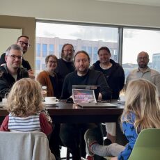Seven adults behind a high table looking into the camera. In front of the table, two young children with their backs to the camera. In the middle of the table and the image alike a sign with the "Homebrew Website Club" logo.