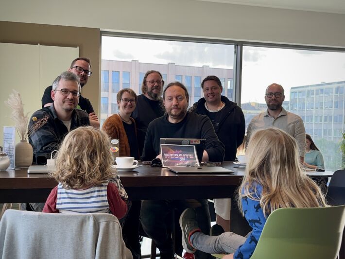 Seven adults behind a high table looking into the camera. In front of the table, two young children with their backs to the camera. In the middle of the table and the image alike a sign with the "Homebrew Website Club" logo.