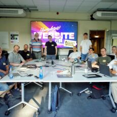 The participants of HWC Nuremberg 07/25 standing around a table. A large Homebrew Website Club logo is in the background.