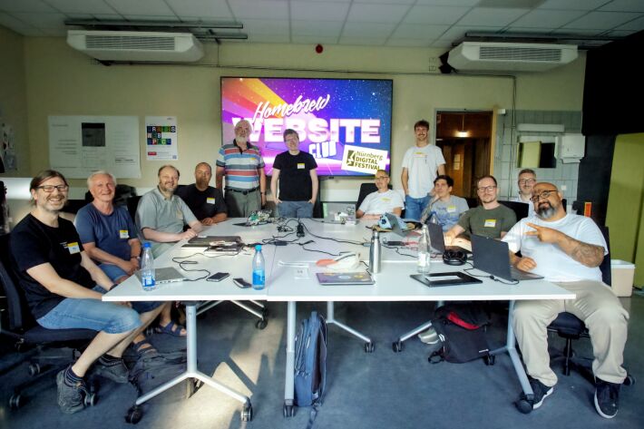 The participants of HWC Nuremberg 07/25 standing around a table. A large Homebrew Website Club logo is in the background.