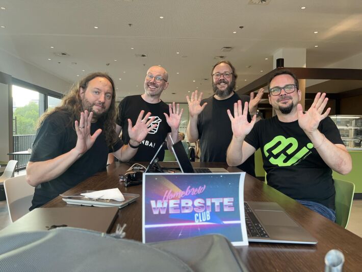 Four smiling participants of the Homebrew Website Club Düsseldorf wave enthusiastically at the camera while sitting around a table with laptops and a "Homebrew Website Club" sign in front. The group is gathered at the café "Xafé" in the KAP1 building (central library) in Düsseldorf, surrounded by a bright, modern interior with large windows.