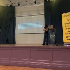 Ana in the stage in front of a slide saying "To help create an alternative to the corporate web, members of the IndieWeb community have built tools that anyone can use on their personal website that helps create the interaction and community building between personal websites."