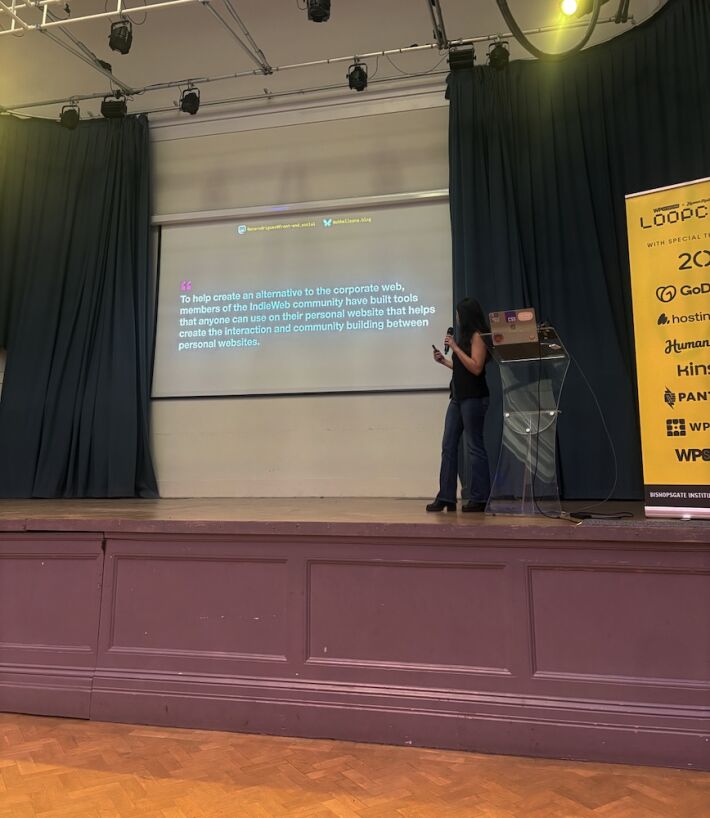 Ana in the stage in front of a slide saying "To help create an alternative to the corporate web, members of the IndieWeb community have built tools that anyone can use on their personal website that helps create the interaction and community building between personal websites."