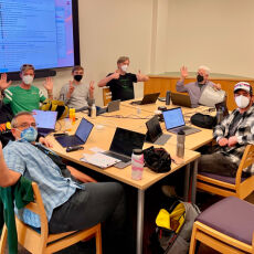 people wearing respiratory masks sitting at a large table inside a meeting room, facing the camera and mostly waving or making thumbs up or peace signs with their hands, in front of a large flat screen on the wall displaying a chat log.