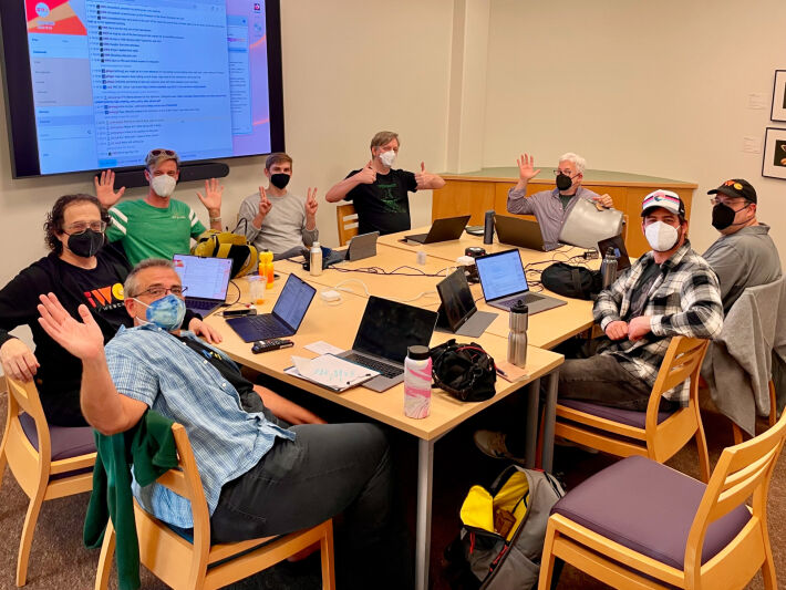 people wearing respiratory masks sitting at a large table inside a meeting room, facing the camera and mostly waving or making thumbs up or peace signs with their hands, in front of a large flat screen on the wall displaying a chat log.