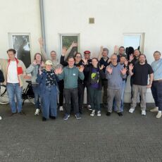 IndieWebCamp Düsseldorf 2026 day 1 one photo with 16 people standing outside in front of a light-colored wall with windows, with most people waving a hand or two in the air