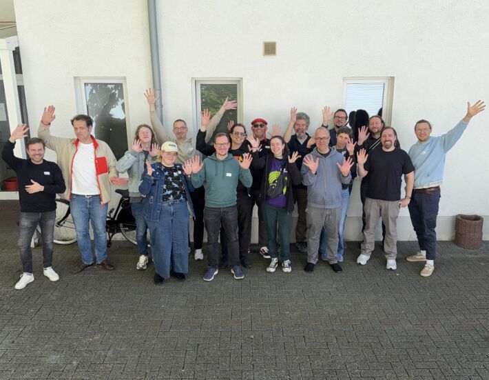 IndieWebCamp Düsseldorf 2026 day 1 one photo with 16 people standing outside in front of a light-colored wall with windows, with most people waving a hand or two in the air
